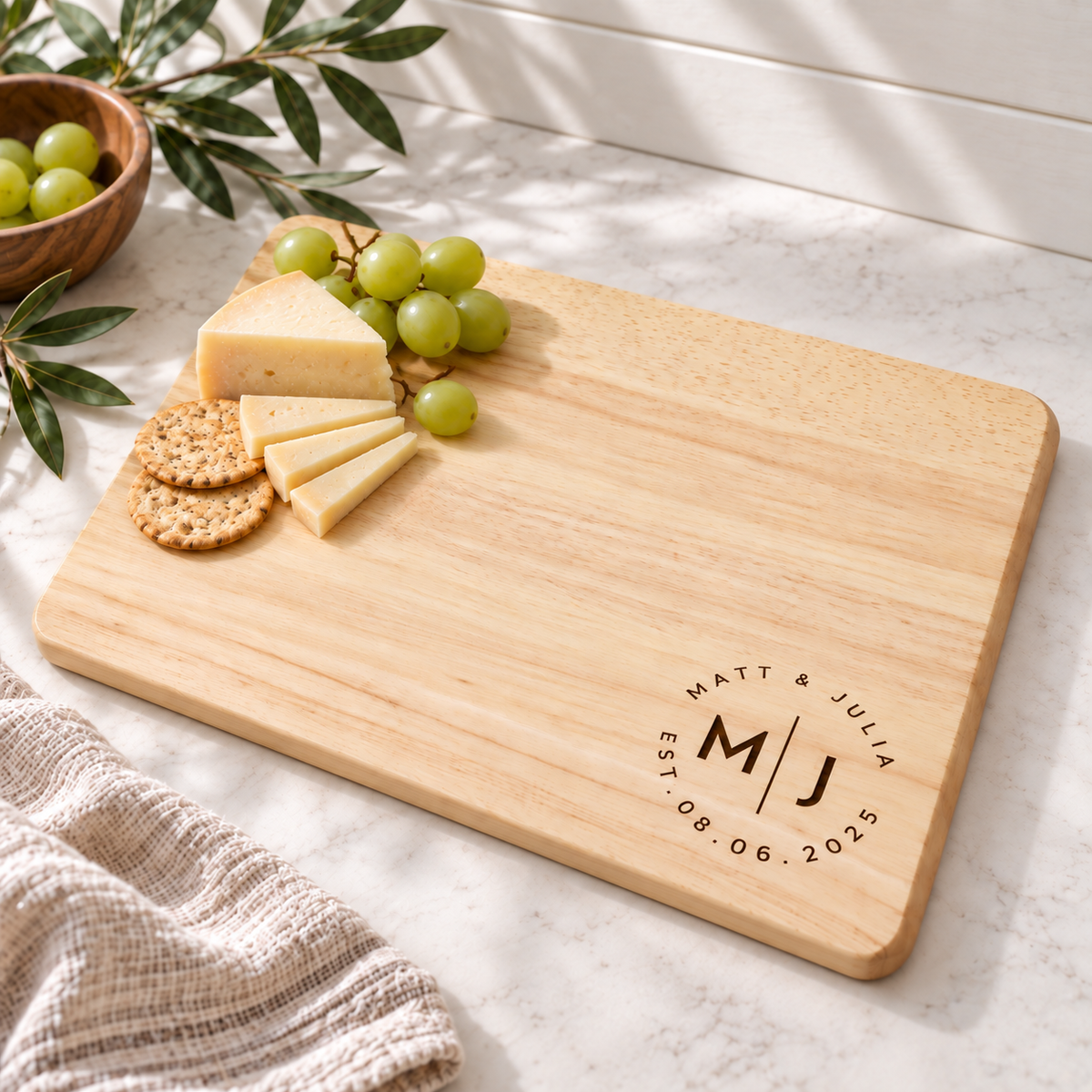 Personalized wooden cheese board with green grapes, cheese wedges, and crackers on a marble surface, photographed in a styled kitchen setting.